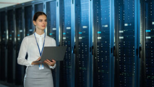 Beautiful Data Center Female IT Technician Walking Through Server Rack Corridor with a Laptop Computer. She is Visually Inspecting Working Server Cabinets.