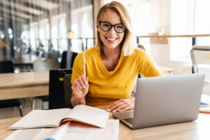 Photo of beautiful happy woman looking at camera while working and sitting at table in open-plan office