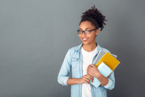 Young african female student isolated on grey wall wearing eyeglasses holding notebooks looking camera smiling