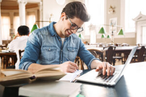 Smiling male student studying at the library, using laptop computer