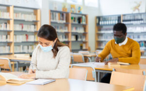 Interested young Latina in medical face mask spending time in library during coronavirus pandemic, reading books and making notes. Compelled precautions concept