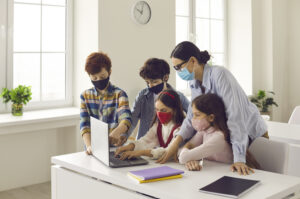 Primary school children and teacher using laptop in classroom. Happy junior students and tutor wearing medical face masks coding video game. Computer lab, technology, basic programming, STEM education