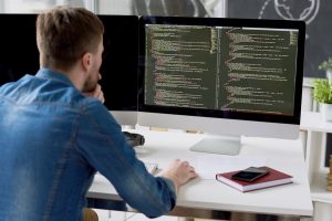 Rear view of concentrated thoughtful male programmer viewing computer language code on computer monitor while working on new program in modern office