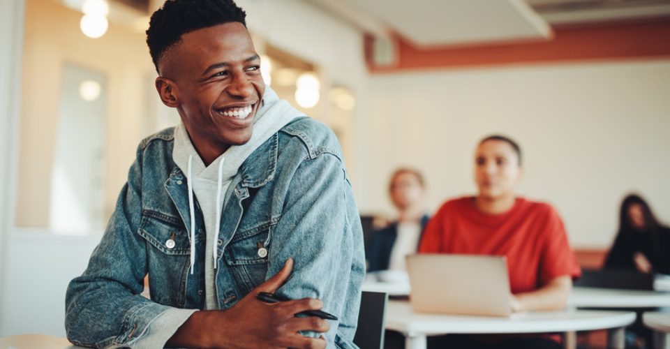 Male student sitting in university classroom looking away and smiling. Man sitting in lecture in high school classroom.