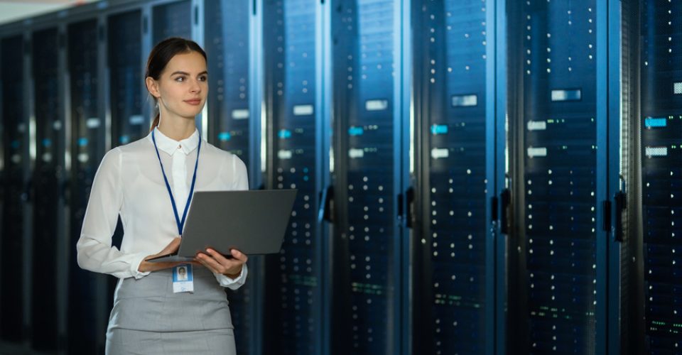 Beautiful Data Center Female IT Technician Walking Through Server Rack Corridor with a Laptop Computer. She is Visually Inspecting Working Server Cabinets.