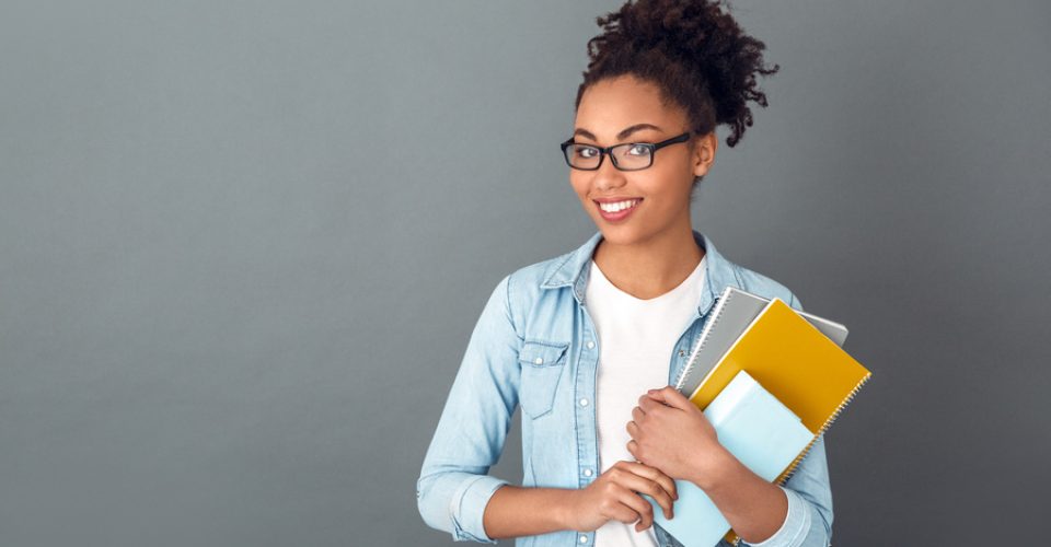 Young african female student isolated on grey wall wearing eyeglasses holding notebooks looking camera smiling