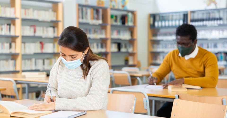 Interested young Latina in medical face mask spending time in library during coronavirus pandemic, reading books and making notes. Compelled precautions concept